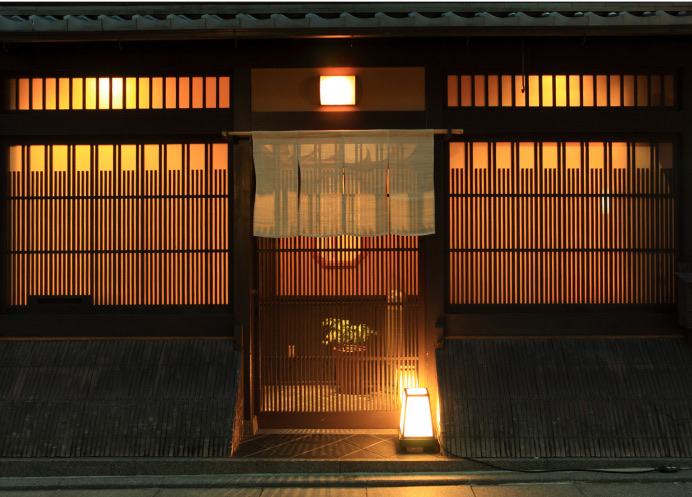 The lattice front of a traditional wooden building at night, lit from within, with a noren curtain over the entrance