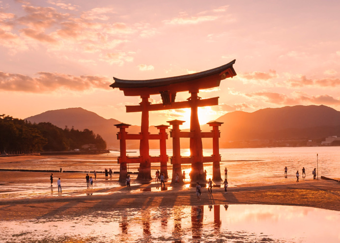 A vermilion torii gate on wet tidal flats at sunset, with people walking around it