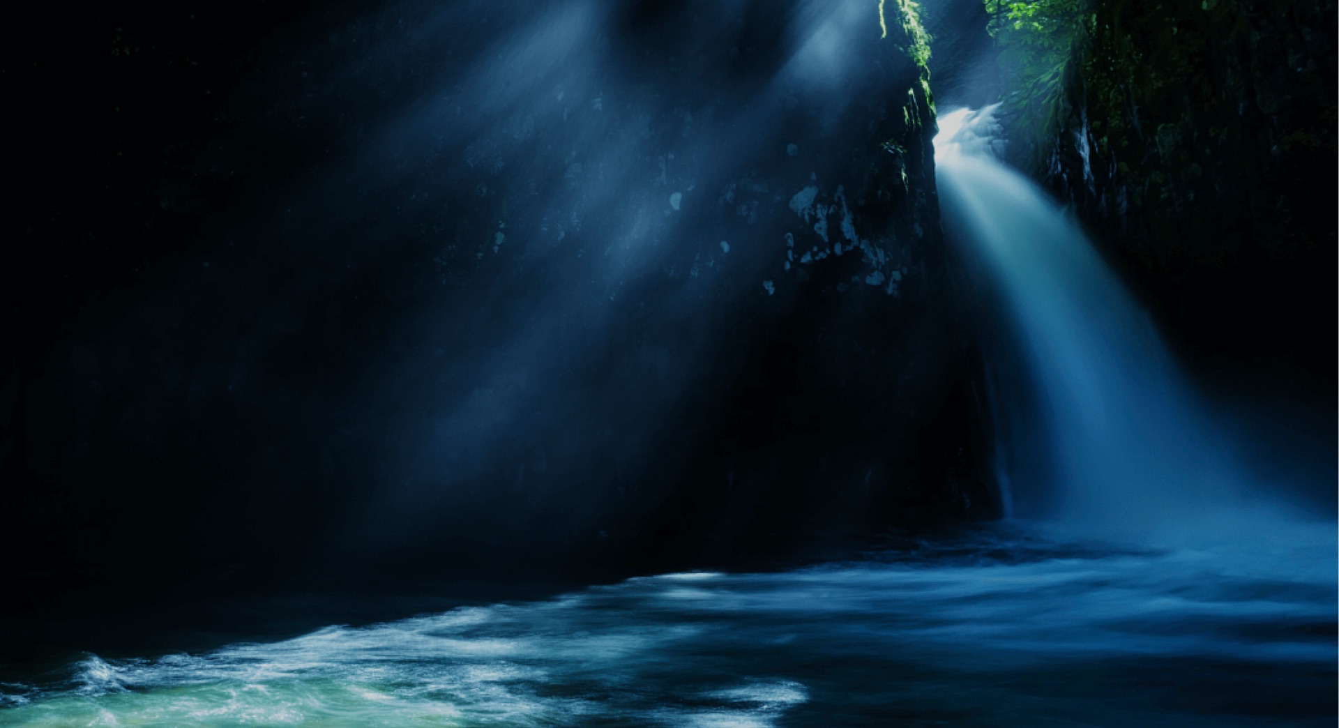 Shafts of light falling through mist beside a waterfall in a dark gorge