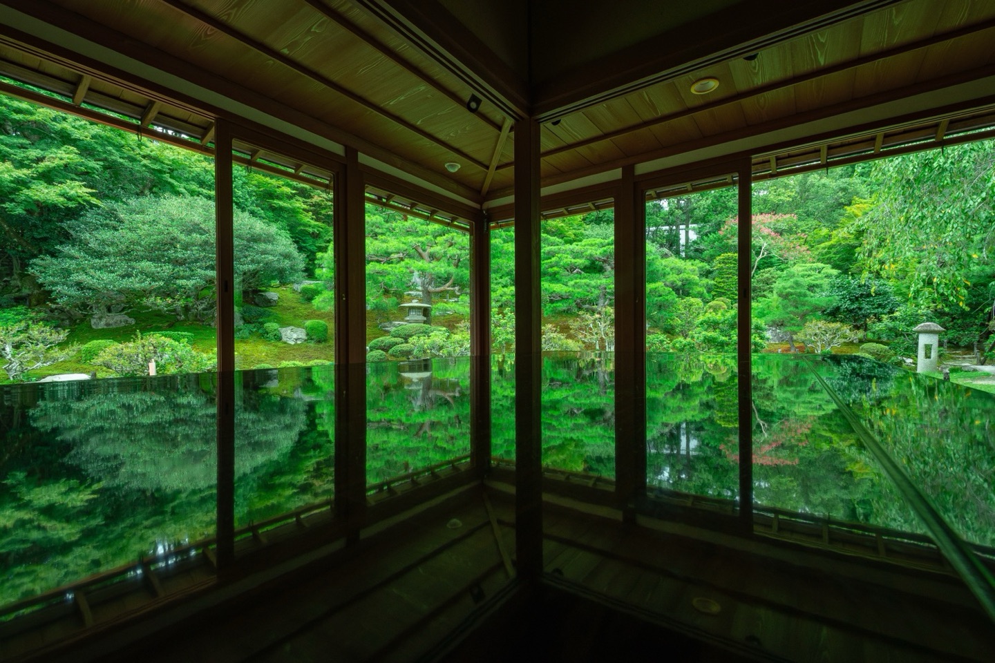 A garden of pines and stone lanterns, seen through the windows of a wooden room and mirrored in a polished surface